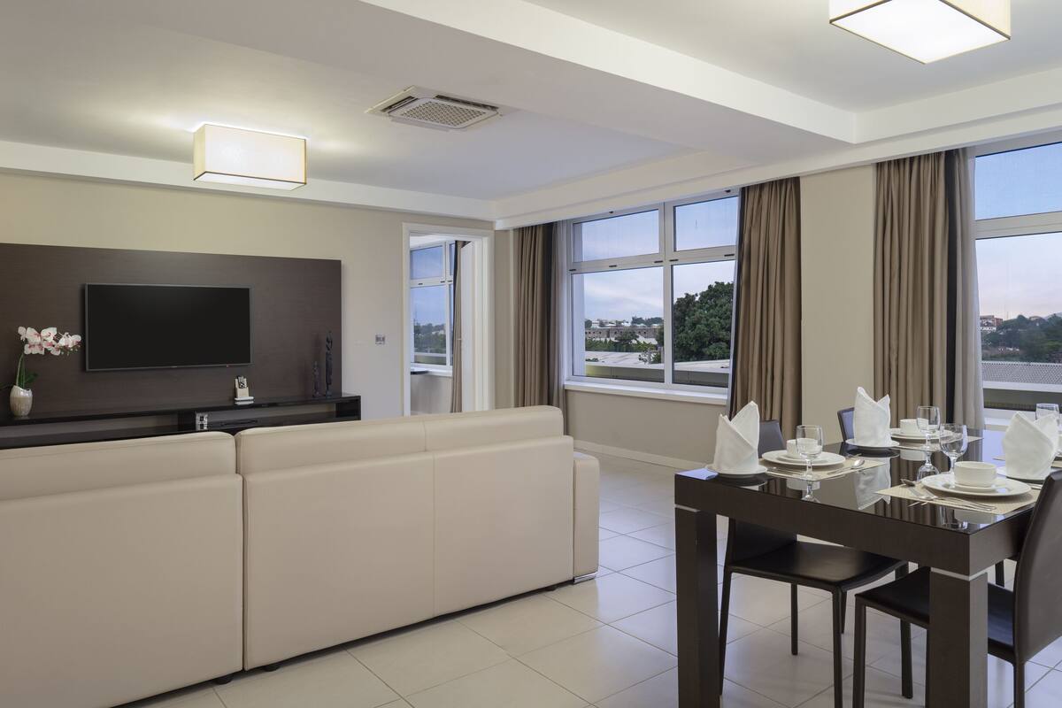Un salon moderne comprenant un canapé, un téléviseur à écran plat et un coin repas aménagé pour quatre personnes. De grandes fenêtres offrent une vue sur la ville.