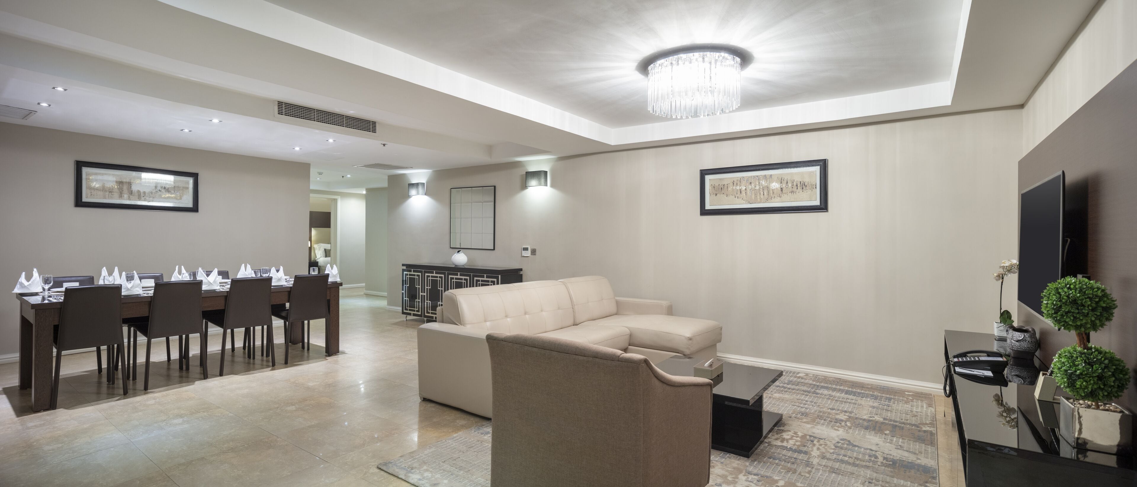A modern living room with a beige sofa, a dining area with a long table and chairs, and a chandelier.