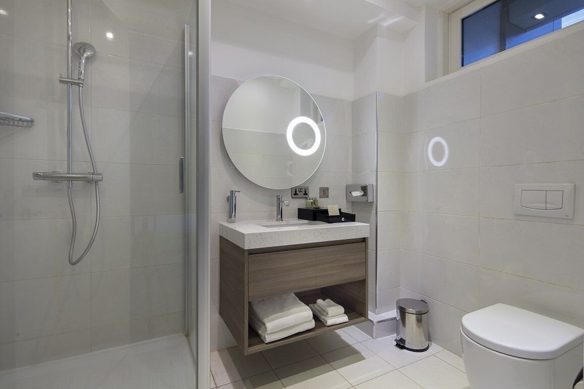 A modern bathroom with a glass-enclosed shower, a round mirror with a light, a white toilet, and a small trash bin.