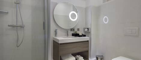 A modern bathroom with a glass-enclosed shower, a round mirror with a light, a white toilet, and a small trash bin.
