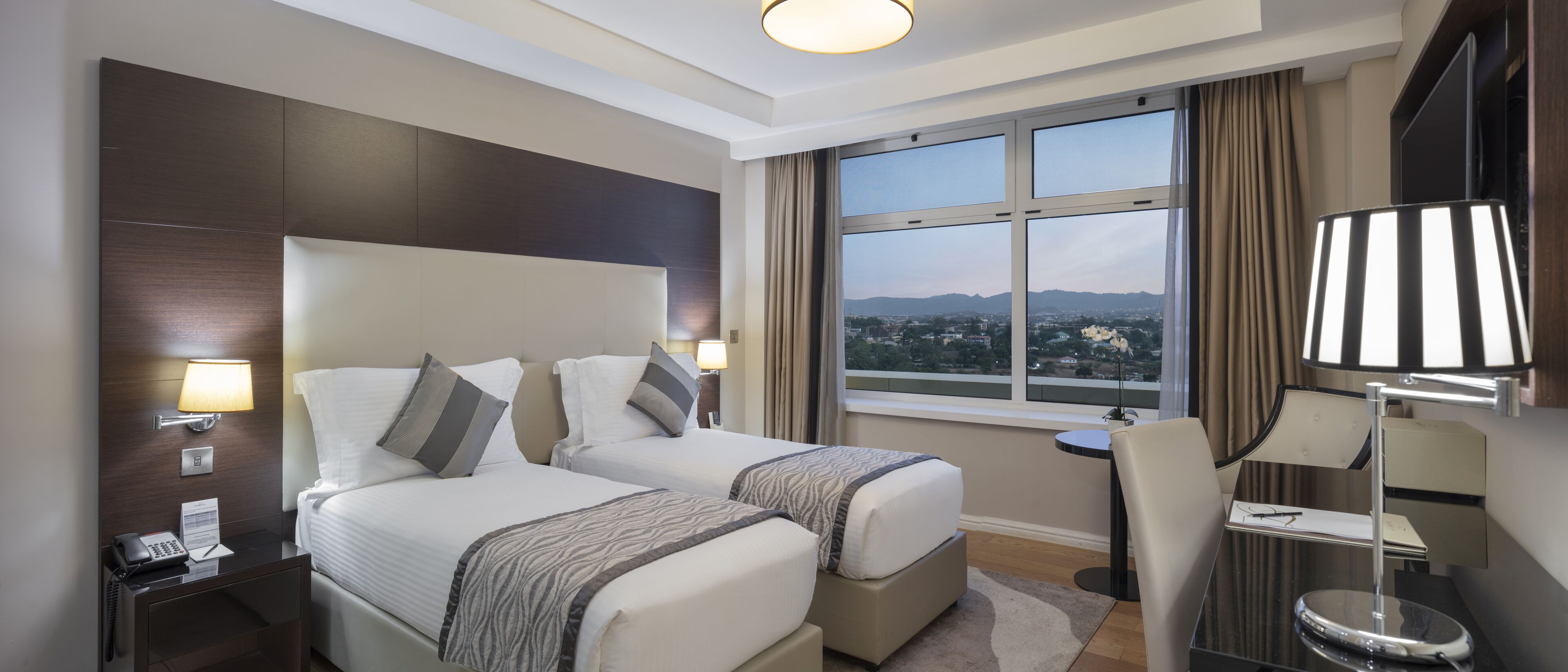 A hotel room with two beds, a desk, a chair, a TV, and a window with a city view.