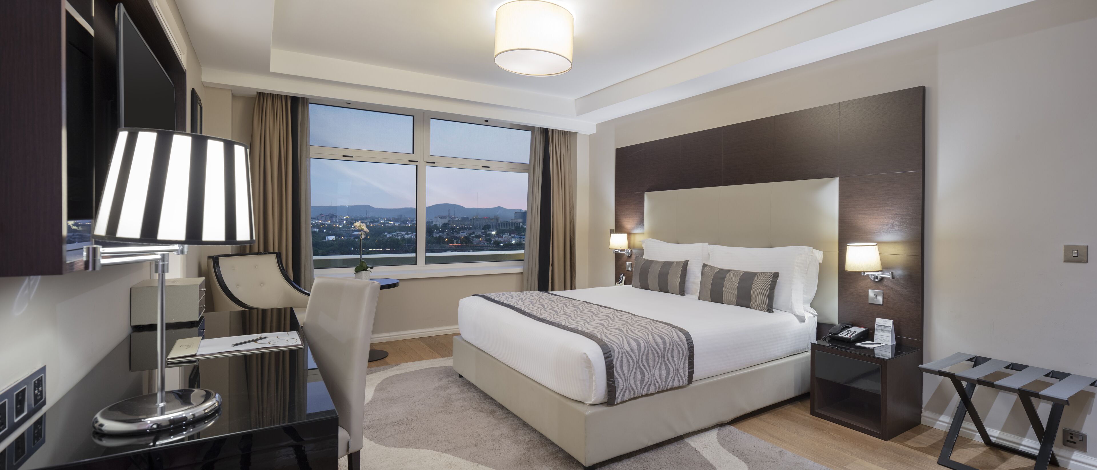A modern hotel room with a large bed, a desk with a lamp, a chair, a nightstand, and a view of the city through large windows.