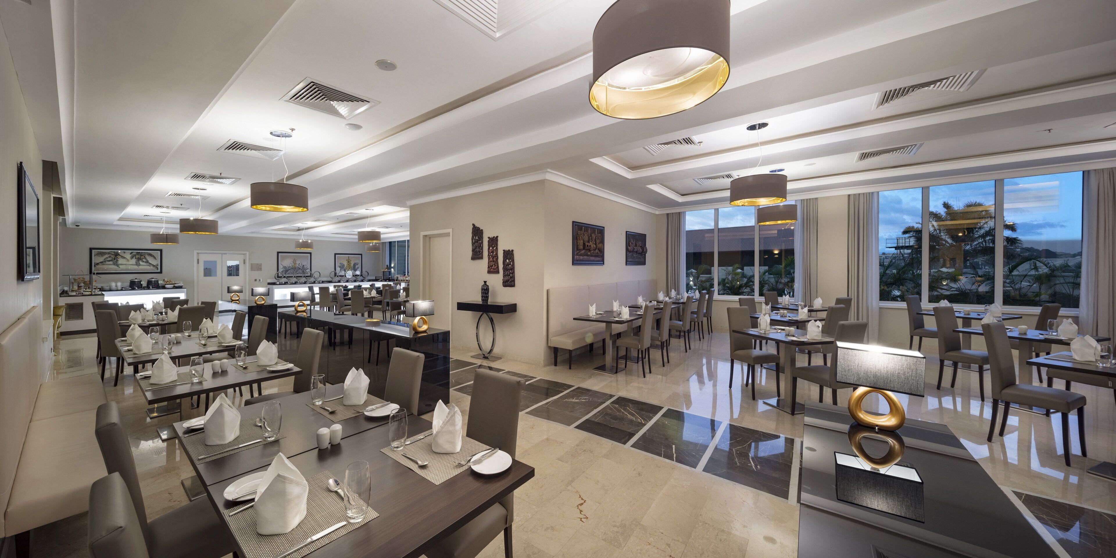 A spacious dining area with multiple tables and chairs, large windows offering a view of the outdoors, and modern pendant lighting.