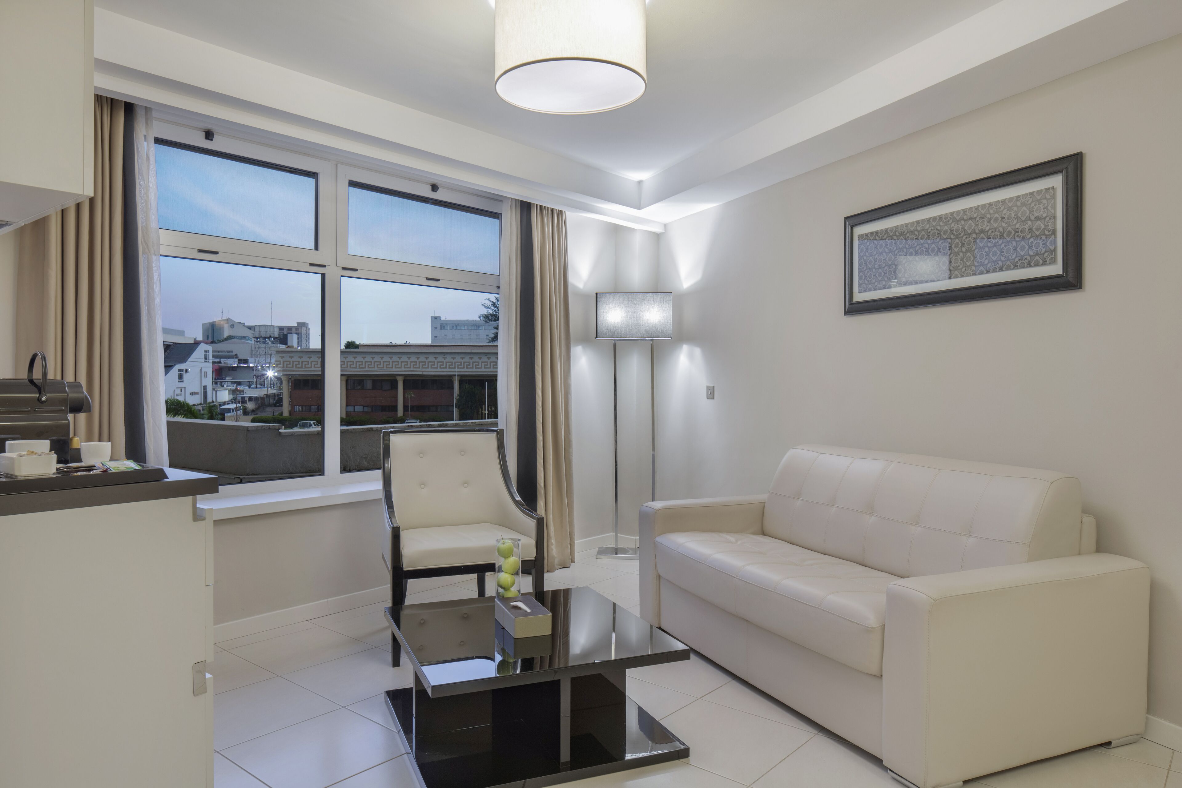 A modern living room with a white sofa, a glass coffee table, a chair, and a floor lamp. There is a large window with curtains, and a framed picture on the wall.