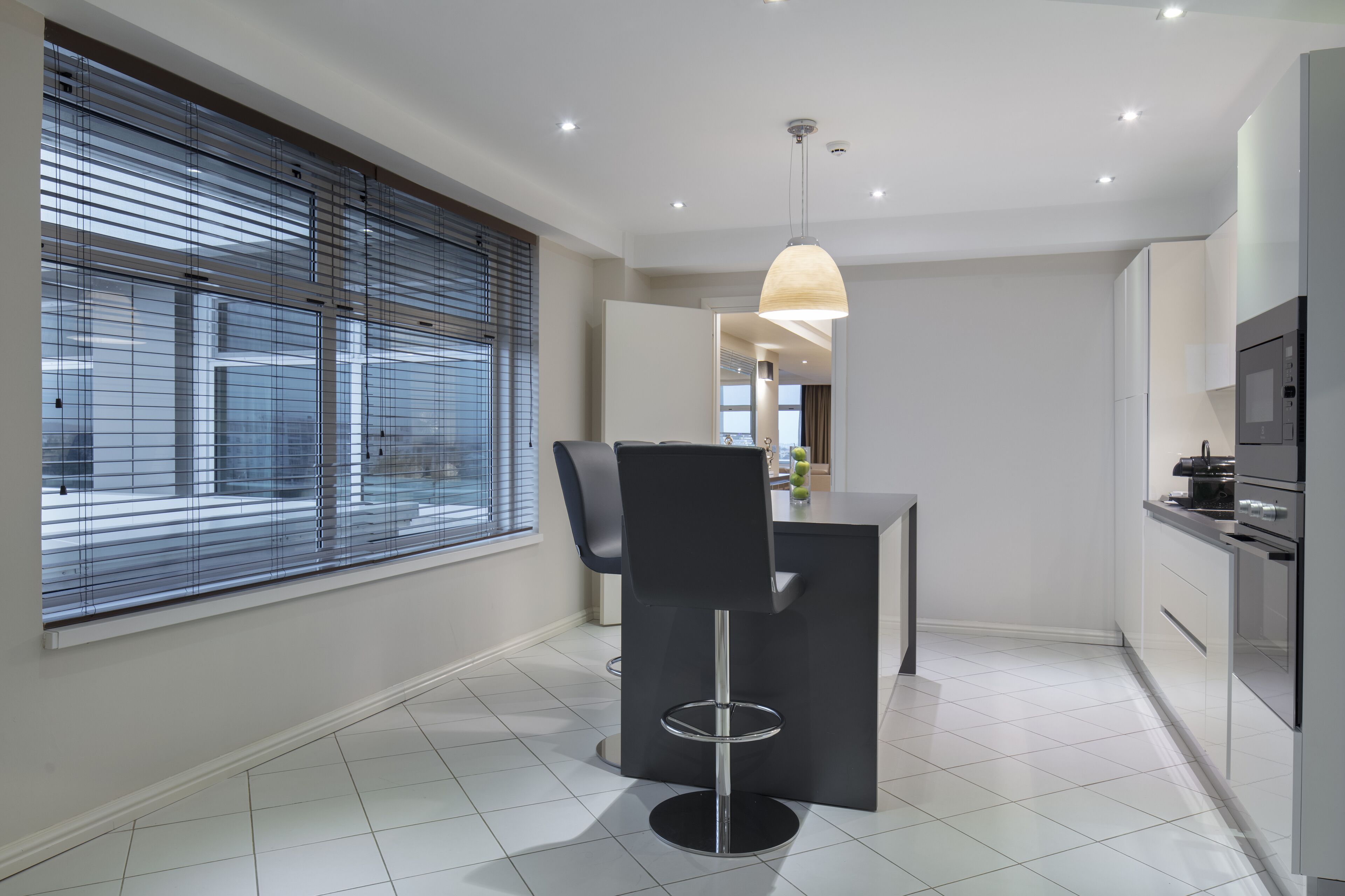 A modern kitchen with a dining area, featuring a large window with blinds, a pendant light, and stainless steel appliances.