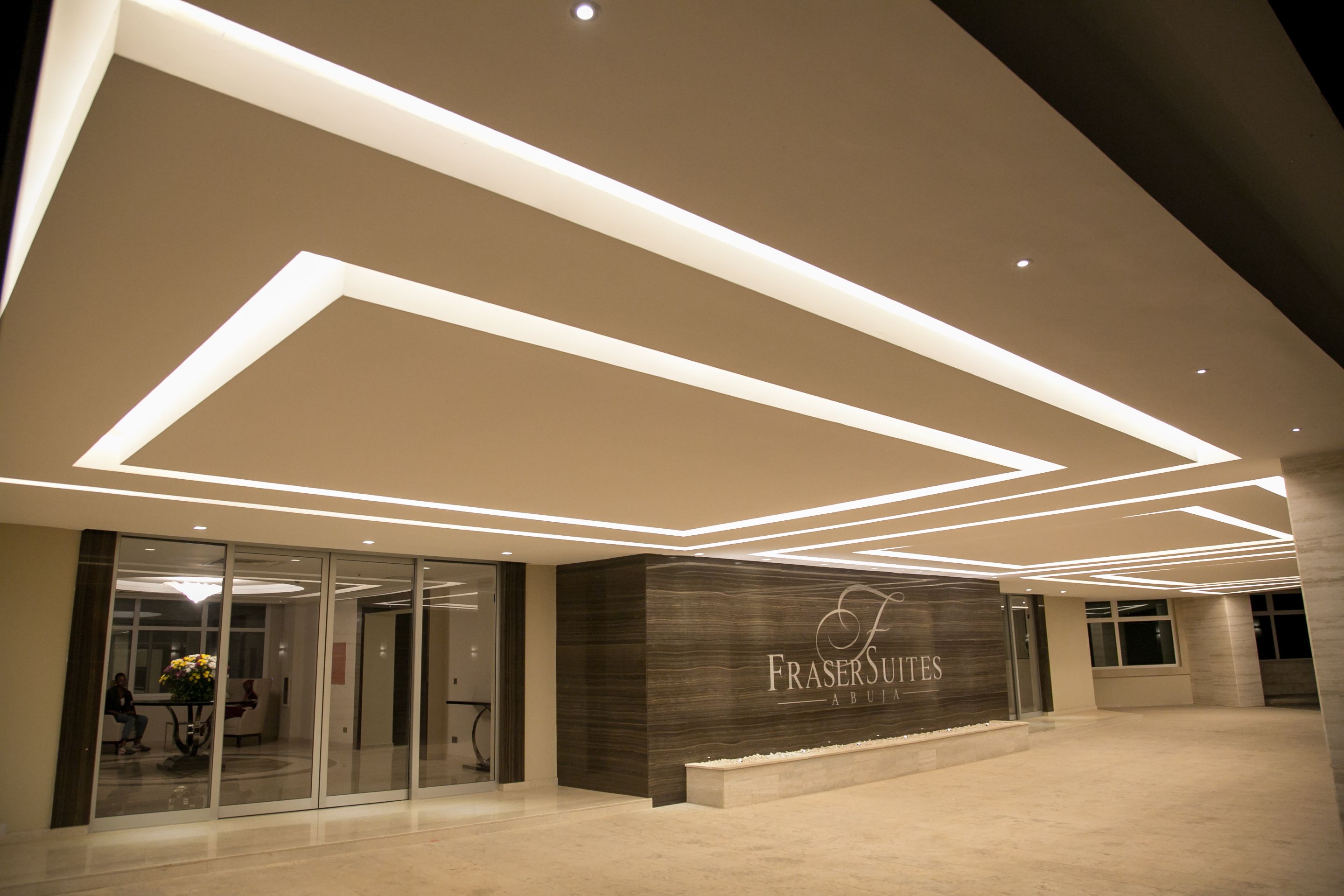 Una entrada de edificio moderna con fachada de vidrio, un mostrador de recepción e iluminación de techo geométrica.