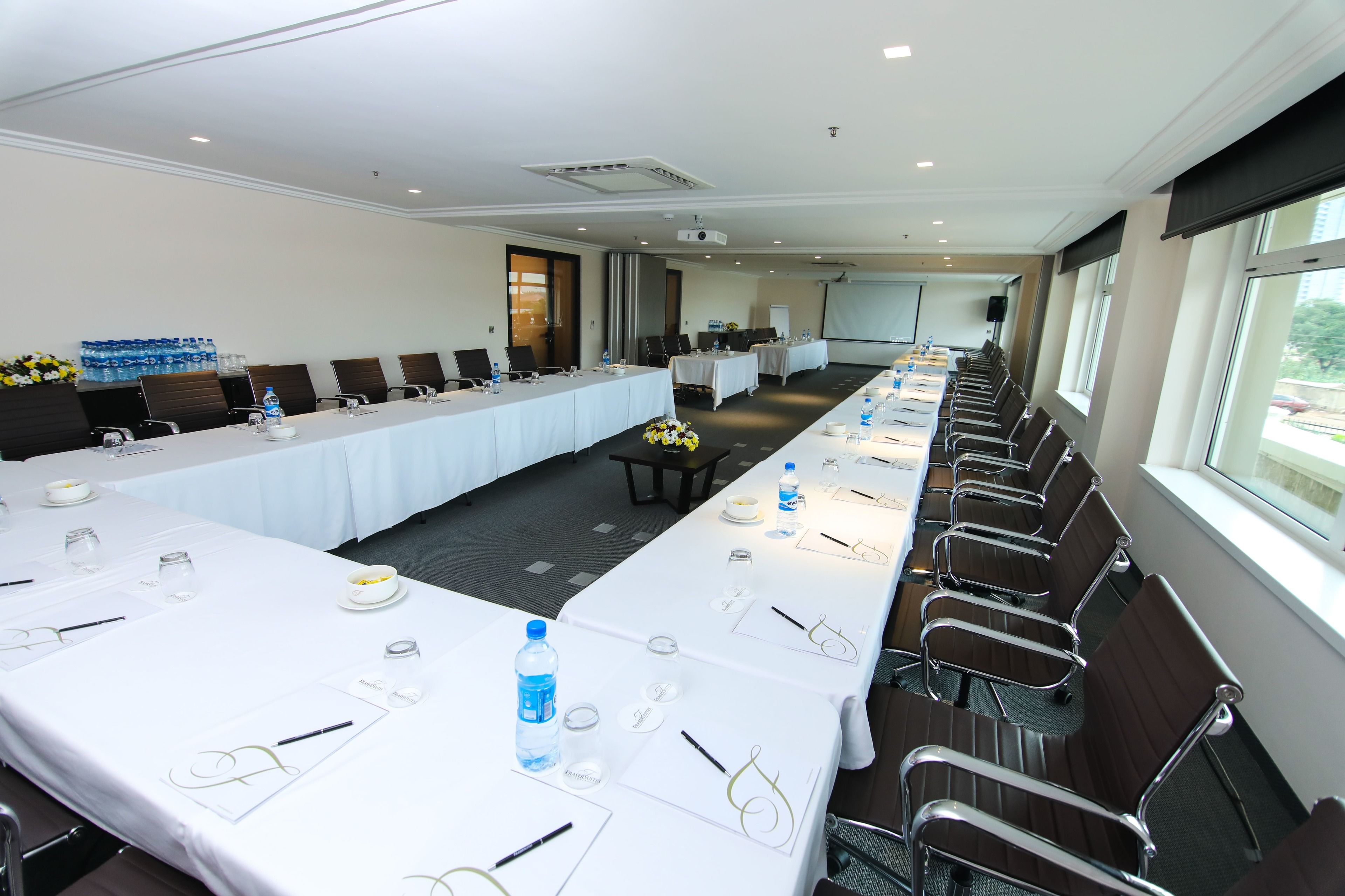 A conference room with long tables, chairs, and a projector screen.