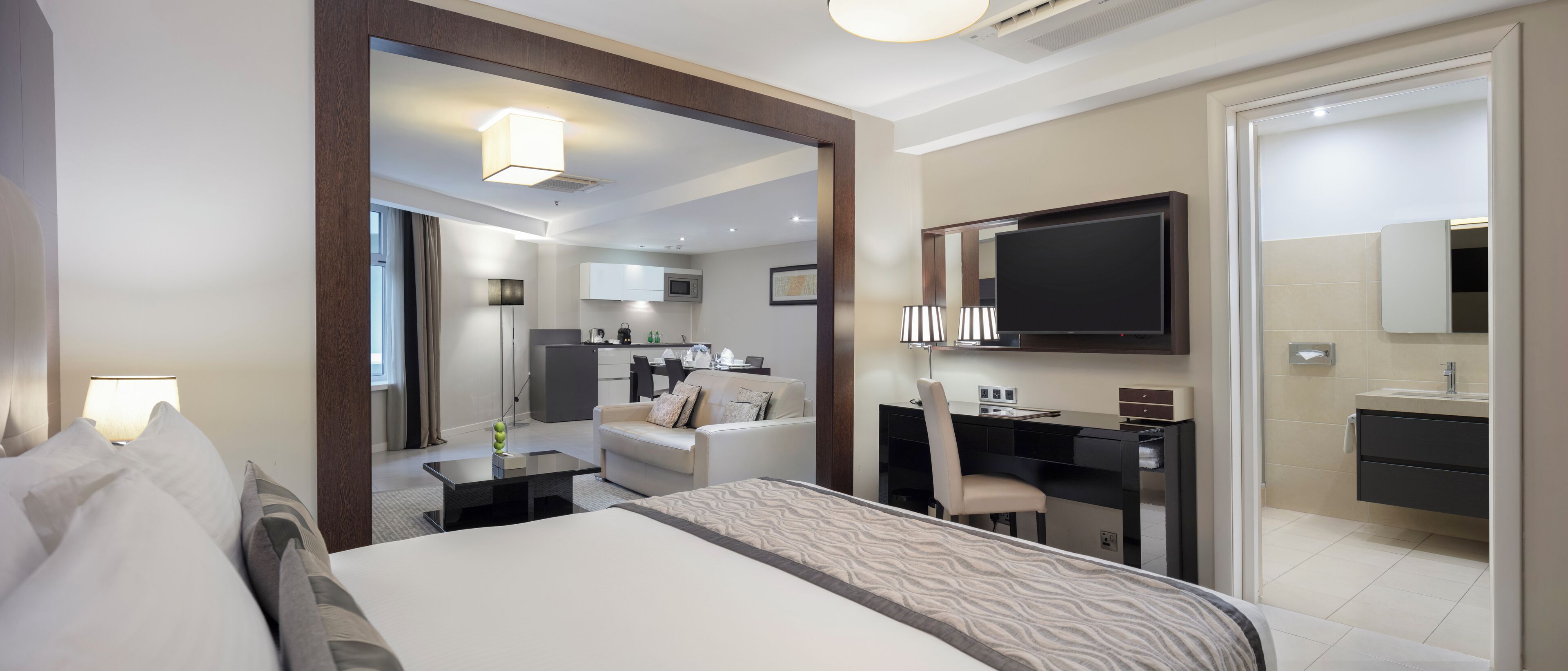 A modern hotel room with a large bed, a desk, a chair, a TV, and a bathroom with a mirror and sink.