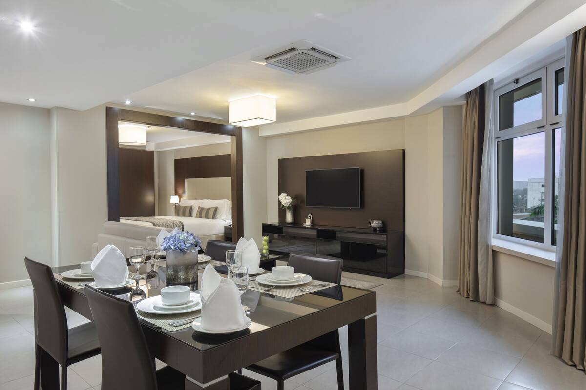 Une chambre d’hôtel moderne dotée d’un coin repas, d’un espace salon avec un téléviseur à écran plat et d’une grande fenêtre avec des rideaux.