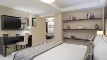 A hotel room with a bed, a desk, a chair, a TV, and shelves with books and decorative items.