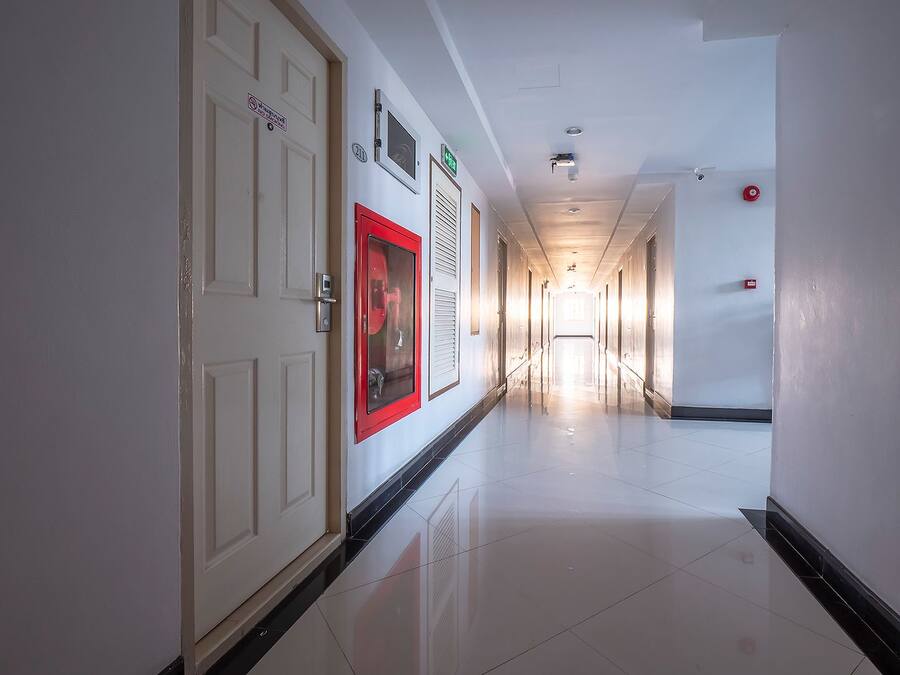 A long, well-lit hallway with doors on both sides, a fire alarm on the right wall, and a fire hose on the left.