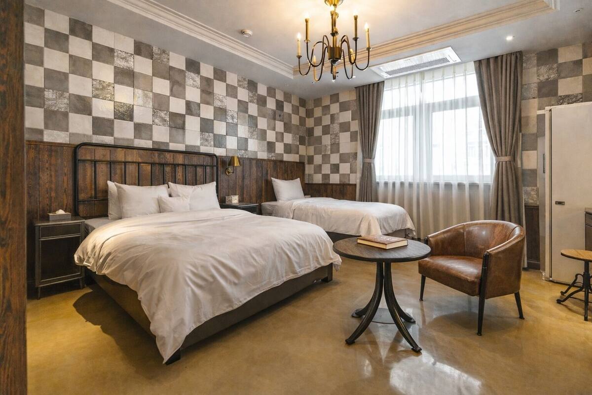 Quarto de hotel com duas camas, um candelabro, cabeceira de madeira, uma mesa redonda, uma poltrona de couro e parede xadrez.