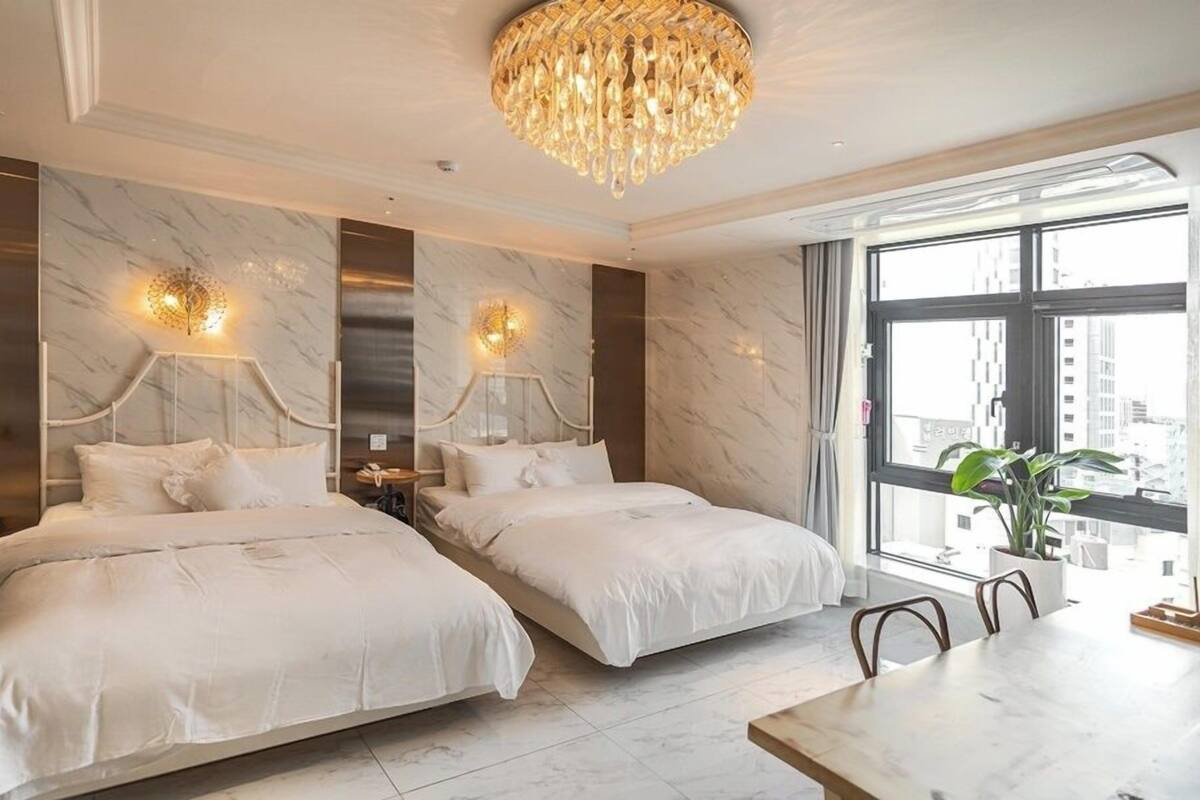 Quarto de hotel com duas camas, um candelabro, uma mesa de jantar e uma janela grande com vista para a cidade.