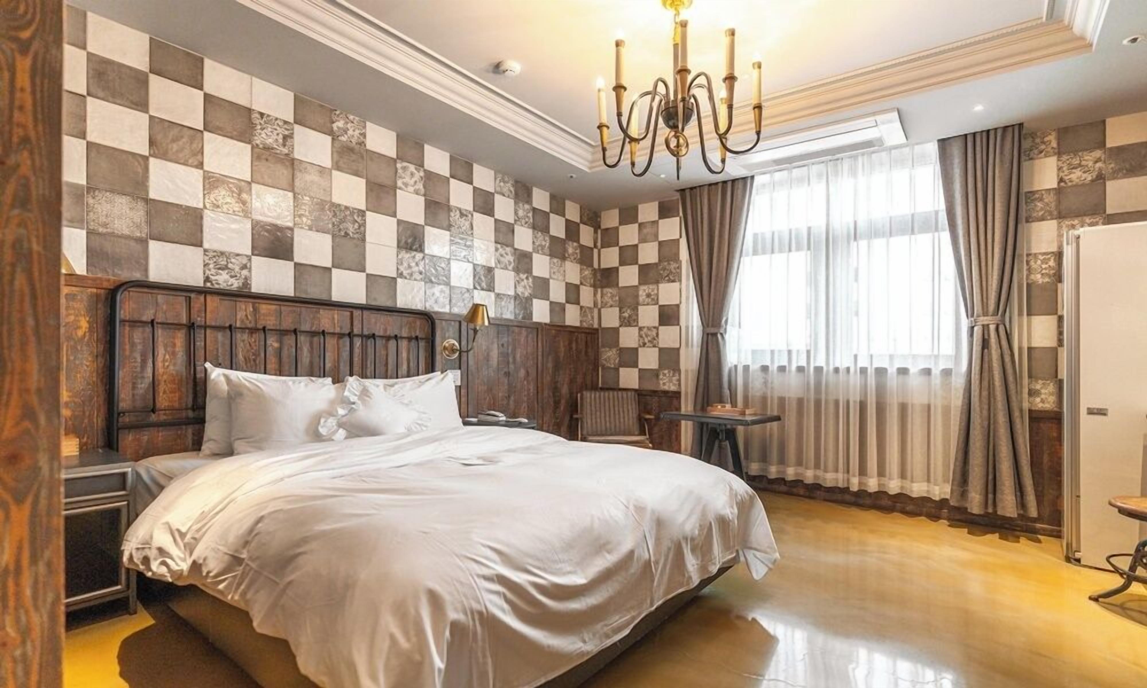A bedroom with a checkered wall, a wooden headboard, a bed with white bedding, a nightstand, a lamp, a window with curtains, and a chandelier.