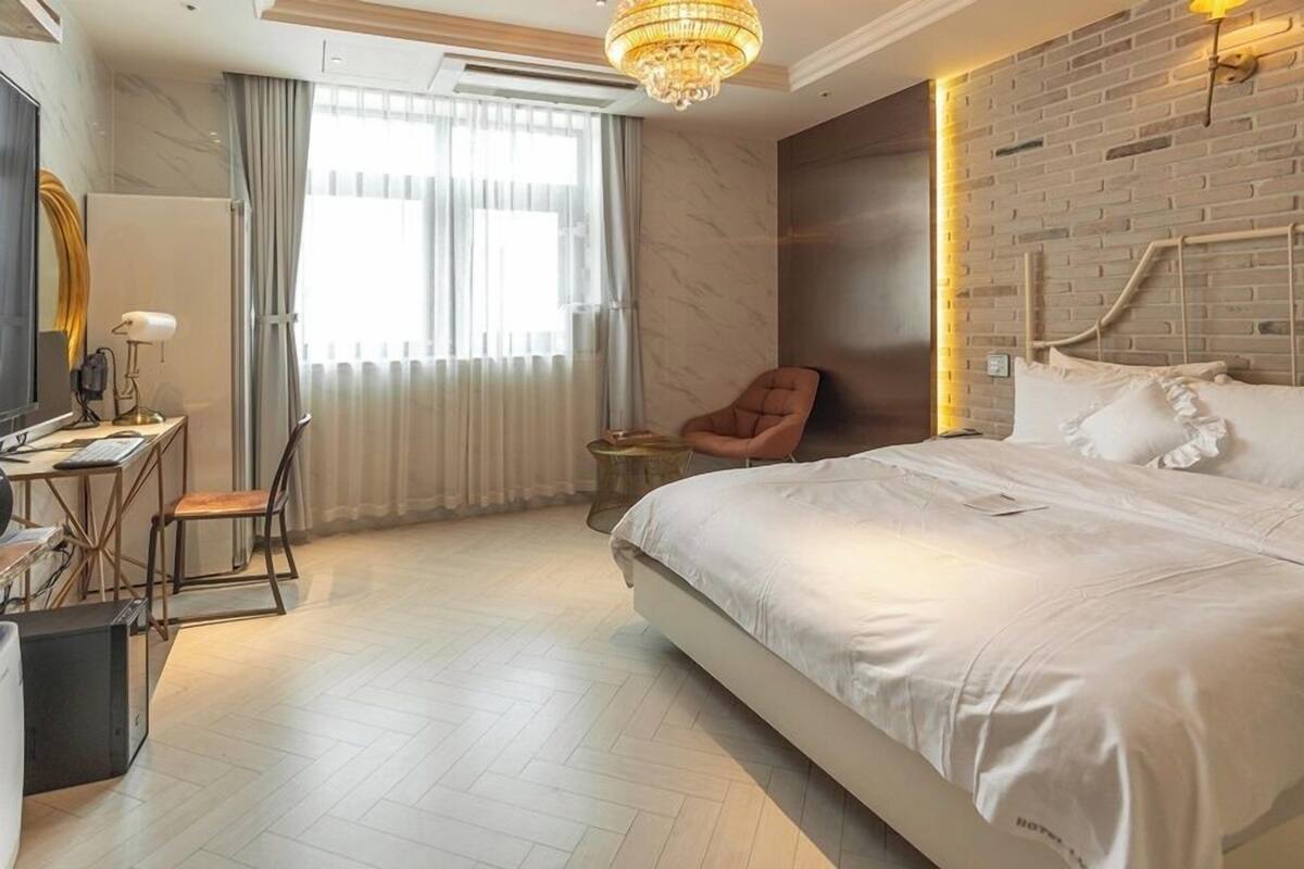 Quarto de hotel moderno com uma cama grande, uma escrivaninha com computador, uma cadeira e uma parede de tijolos.