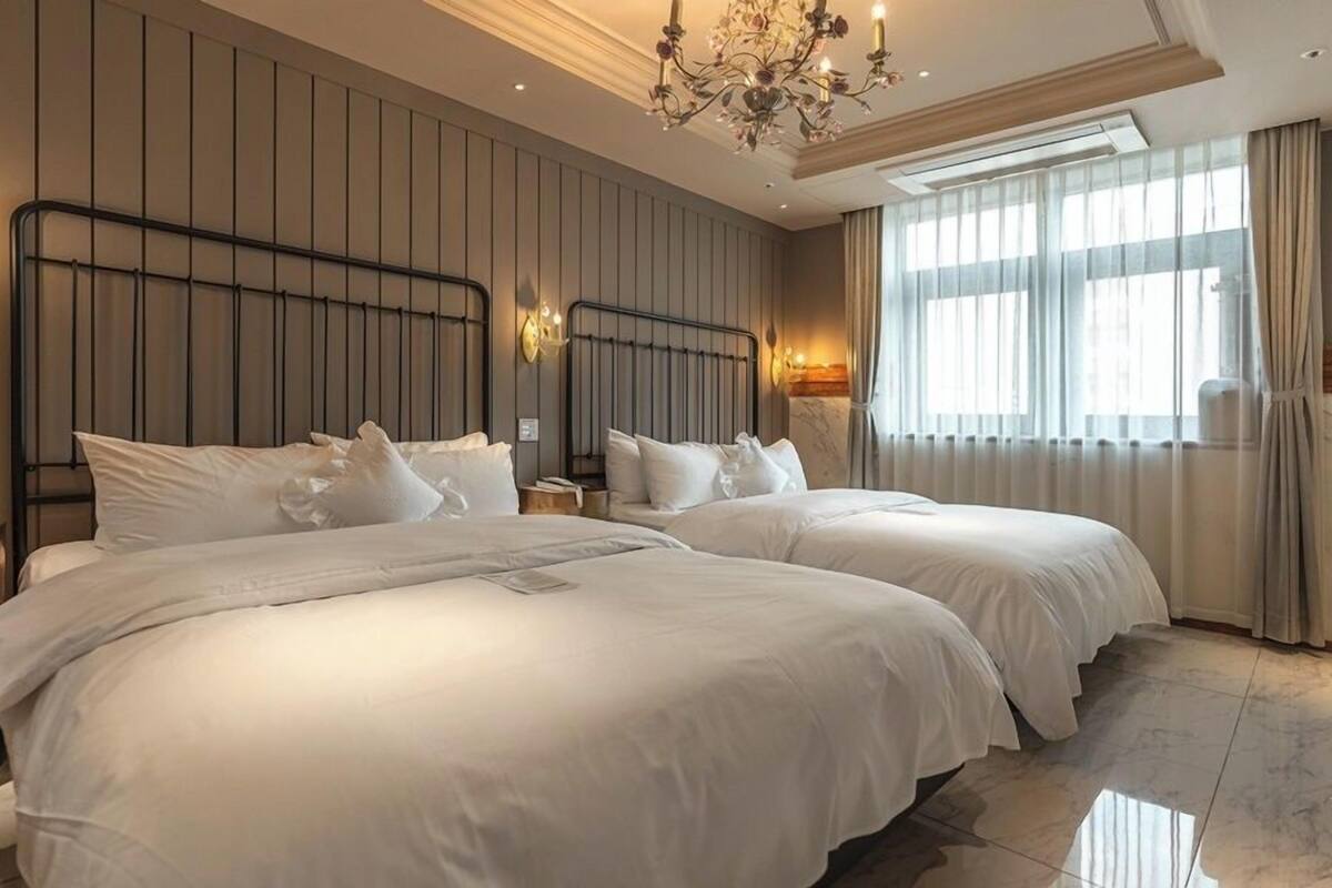 Quarto de hotel com duas camas, um candelabro, uma janela grande com cortinas e um aplique na parede.