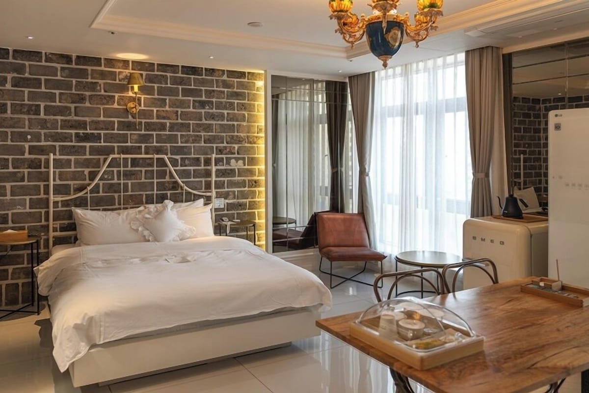 A hotel room with a brick wall, a bed, a dining table, a chair, a refrigerator, and a chandelier.