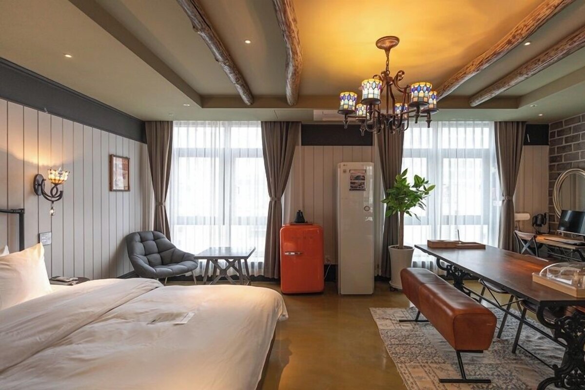 A hotel room with a bed, a desk, a chair, a bench, a TV, a mirror, and a chandelier.