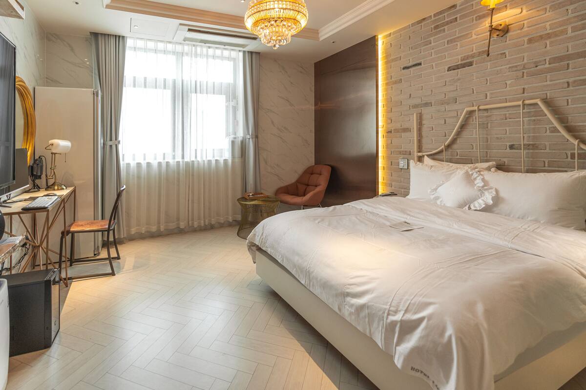 Quarto de hotel moderno com uma cama grande, uma escrivaninha com computador, uma cadeira e um candelabro.