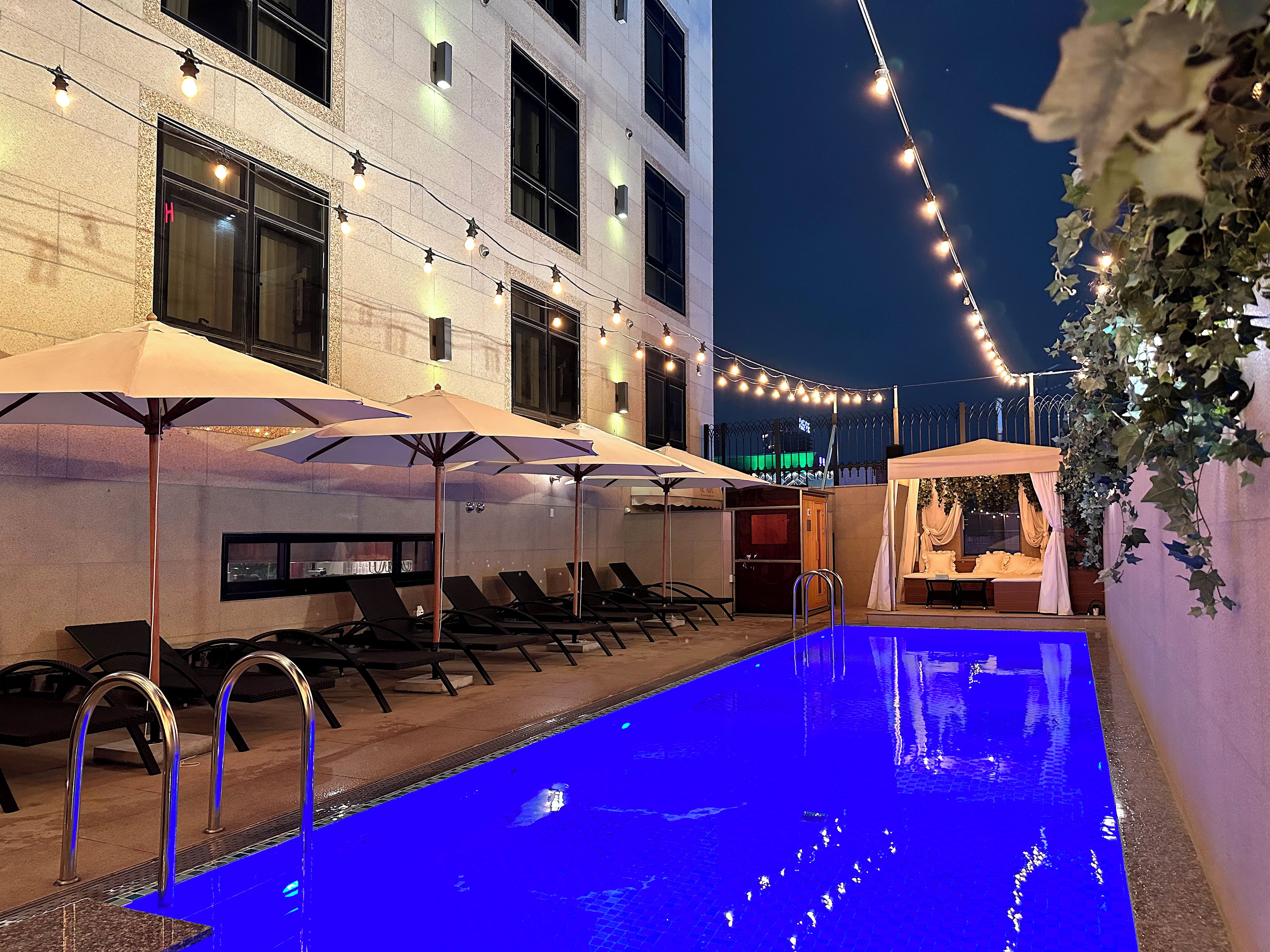 A pool area with lounge chairs and umbrellas, a covered seating area, and string lights.