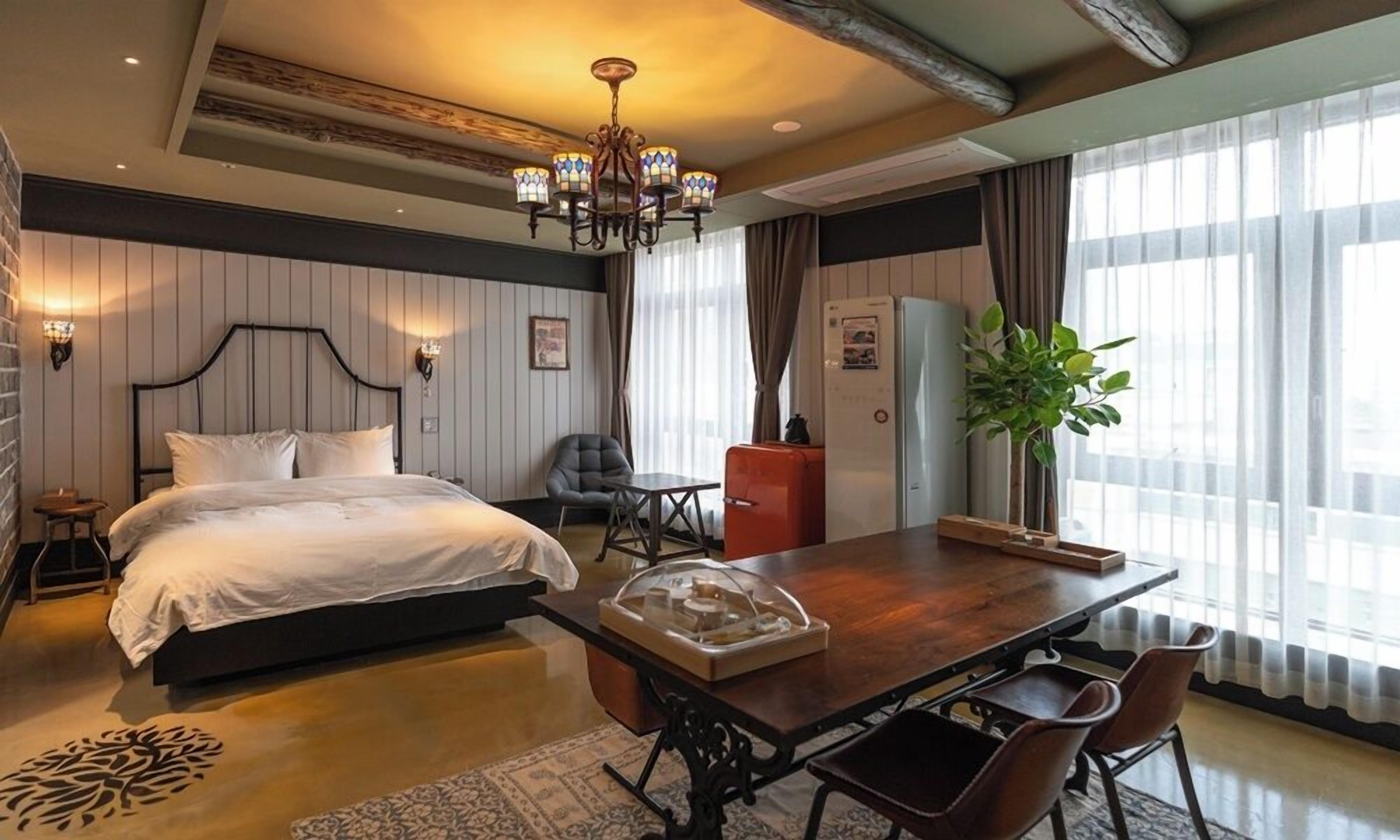A hotel room with a bed, a dining table with chairs, a chandelier, and a large window with curtains.