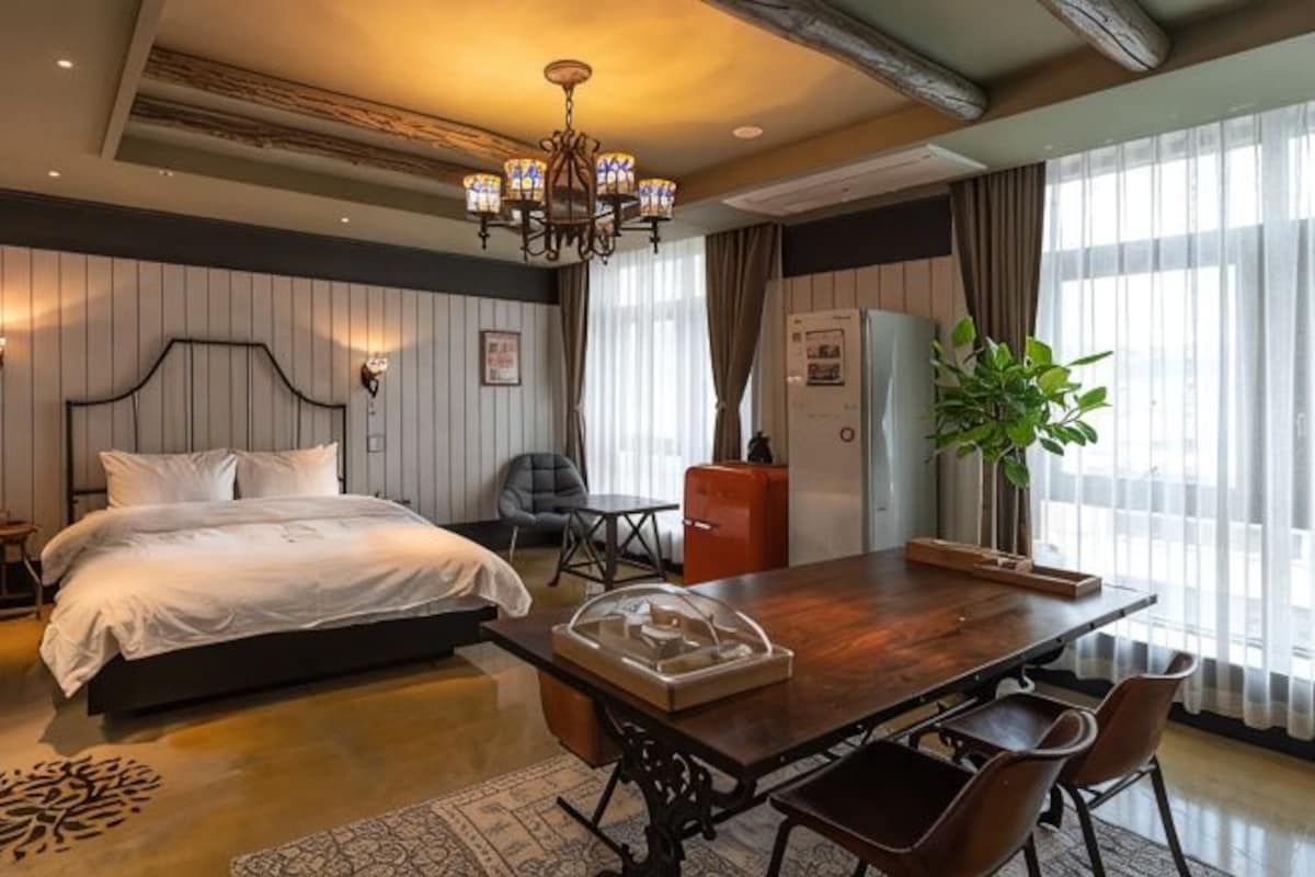 A hotel room with a bed, a dining table, chairs, a chandelier, a plant, and a large window.