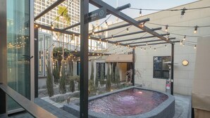 Un espace piscine aménagé sur le toit, avec un bassin en pierre, entouré de murs en verre et agrémenté d’une pergola illuminée de guirlandes lumineuses.