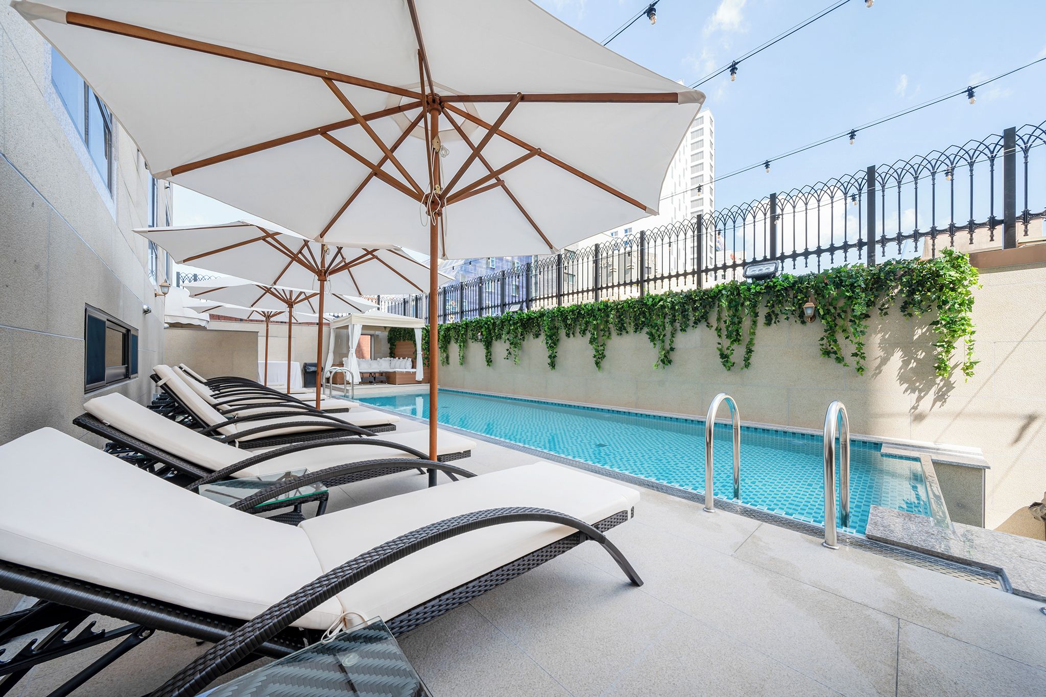 Un área de piscina en la azotea con sillones de descanso y sombrillas, rodeada por una cerca alta y un edificio cubierto de hiedra.