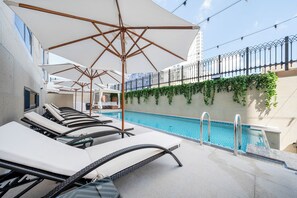 A rooftop pool area with lounge chairs and umbrellas, surrounded by a high fence and a building with ivy.
