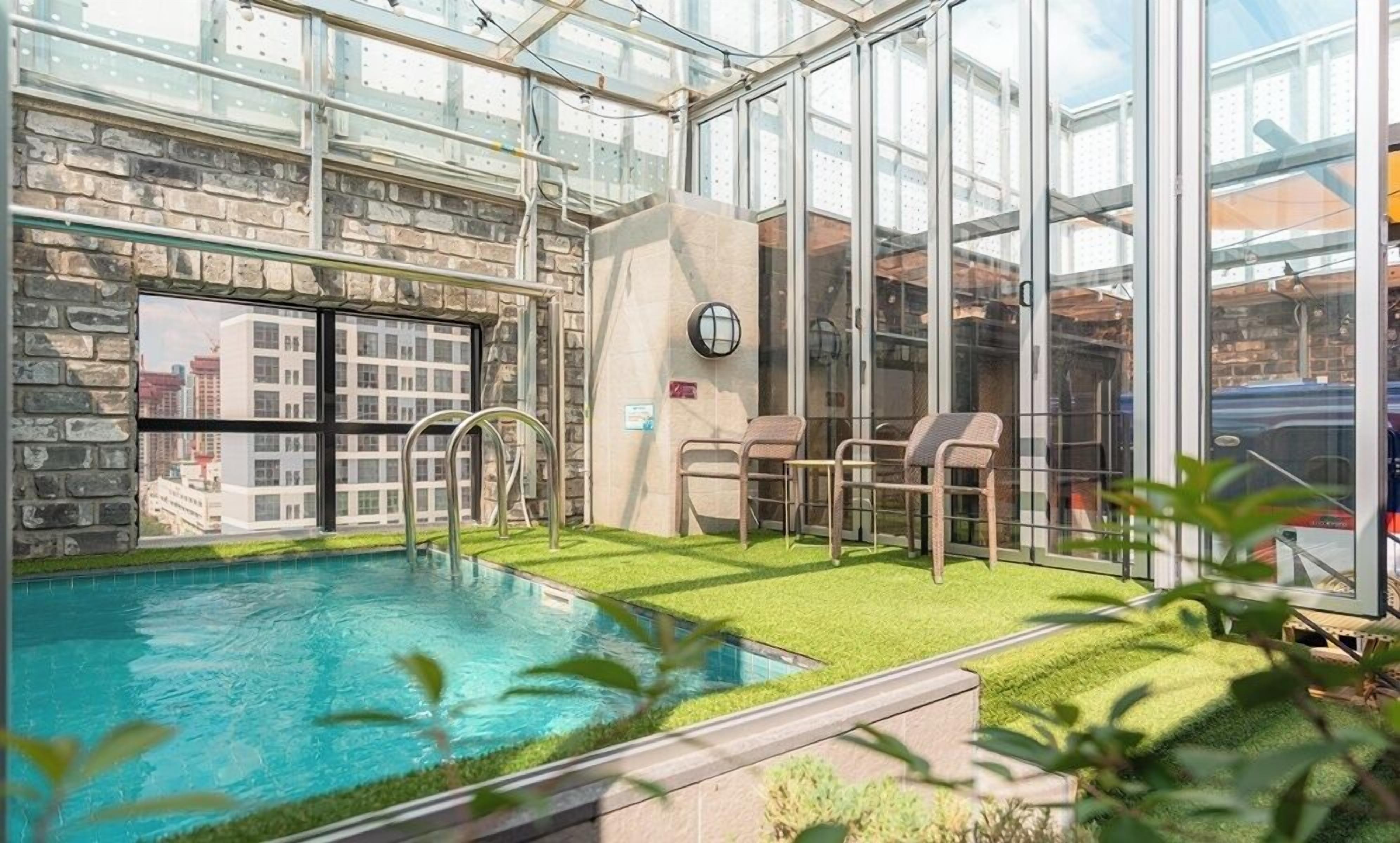 A rooftop pool with glass walls, a stone wall, and two chairs.