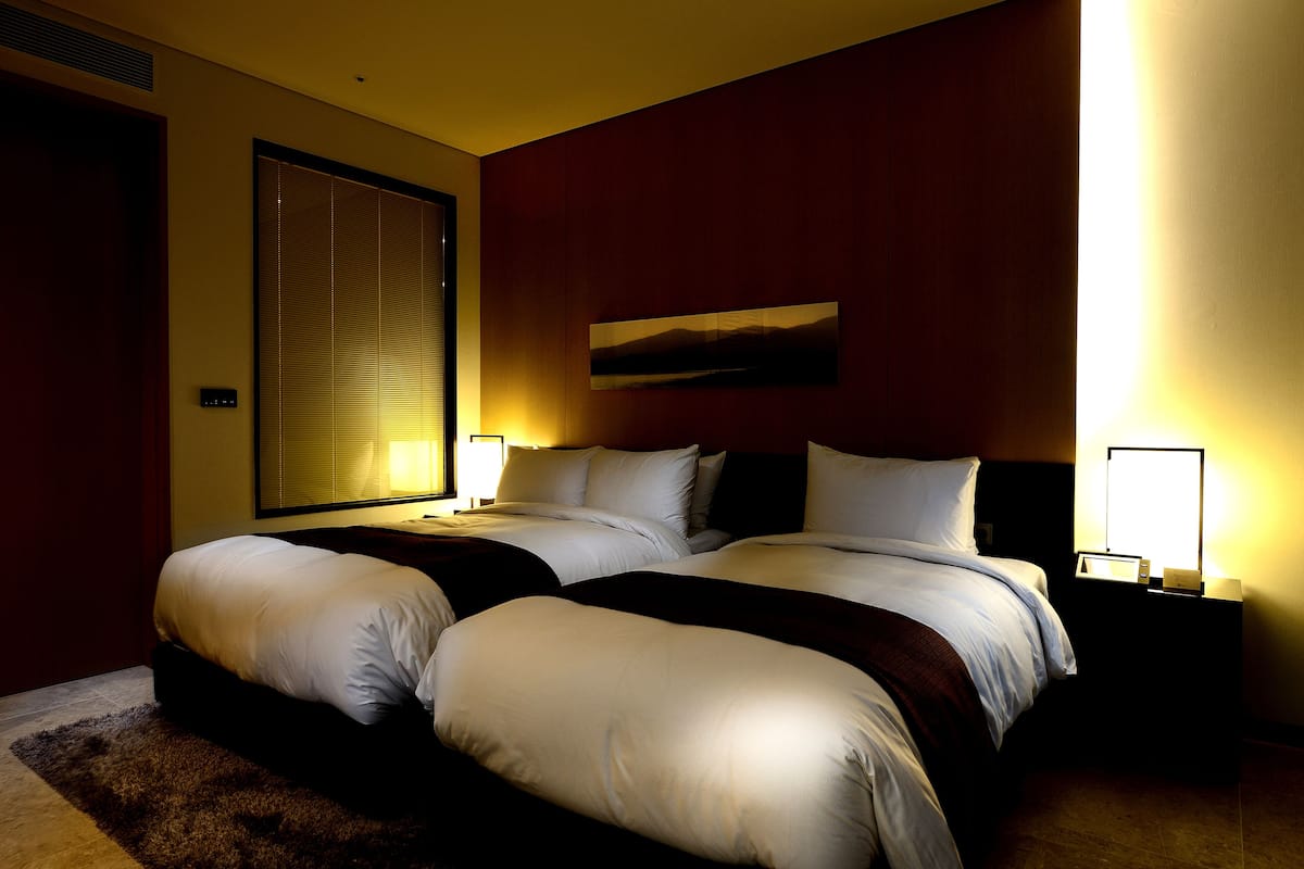 A hotel room with two beds, a large window with blinds, a painting on the wall, and bedside lamps.