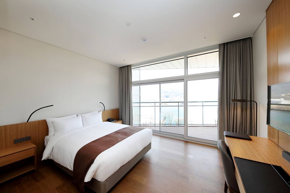A modern hotel room with a large bed, a desk, a TV, and a view of the city.