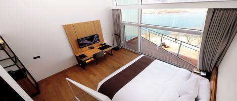 A hotel room with a bed, a desk with a TV, a balcony with a view of the water, and a wooden floor.