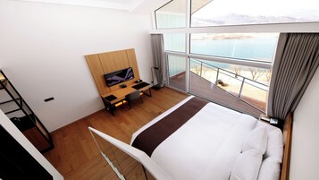 A hotel room with a bed, a desk with a TV, a balcony with a view of the water, and a wooden floor.