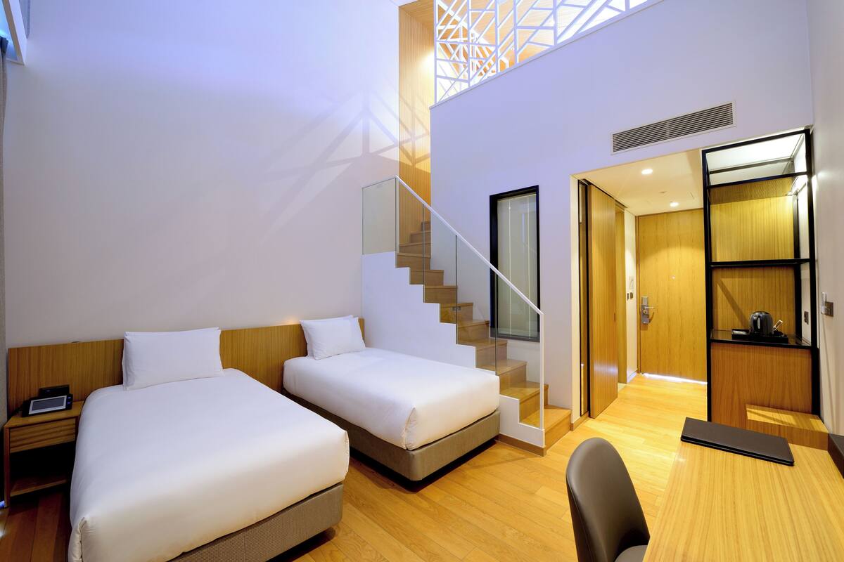 A modern hotel room with two beds, a staircase, a desk, and a kitchenette.