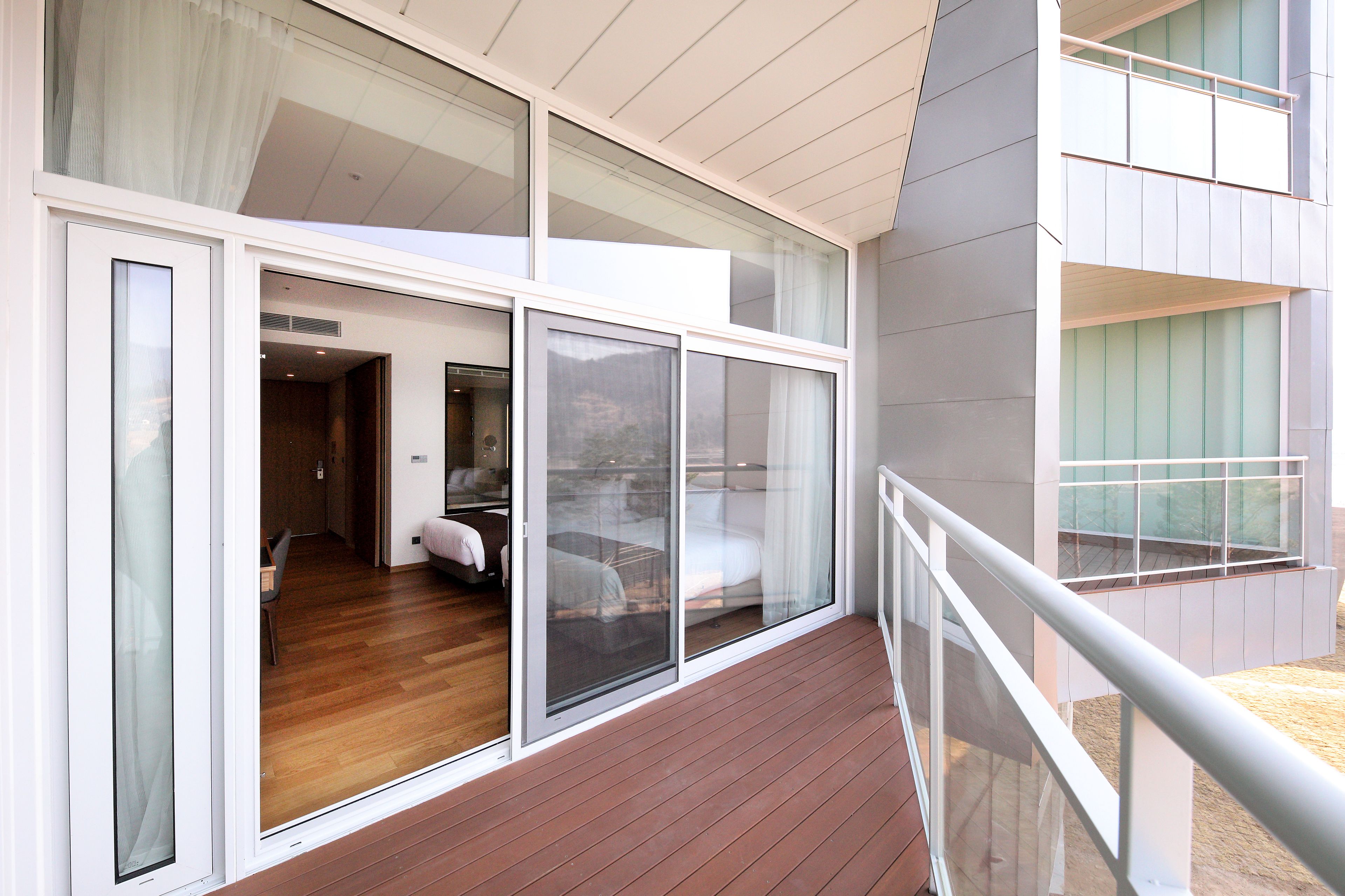 A modern hotel room with a wooden floor, large sliding glass doors, and a balcony with a metal railing.