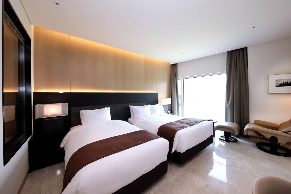 A hotel room with two beds, a sofa, a chair, a large window, and a framed picture on the wall.