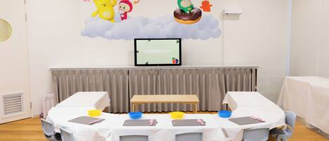 A room with a round table, chairs, a television screen, and a wall with animated characters.