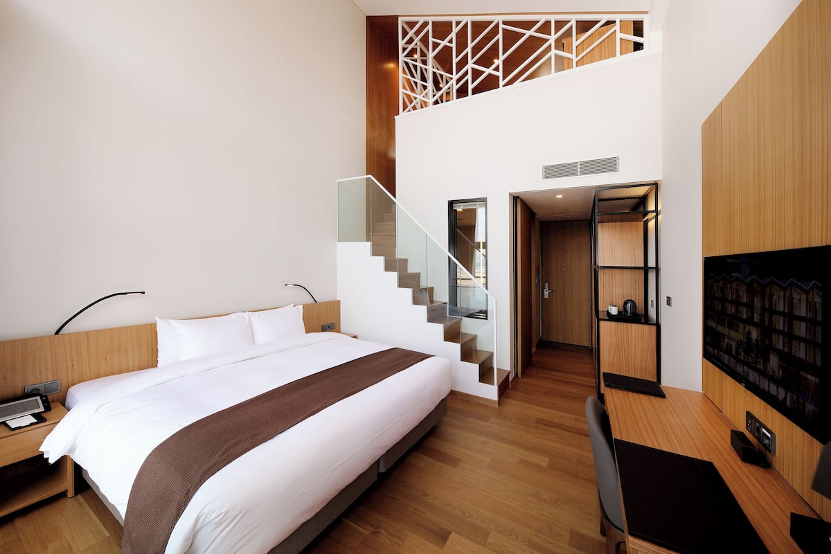 A modern hotel room with a large bed, a desk, a flat-screen TV, a bookshelf, and a staircase leading to an upper level.