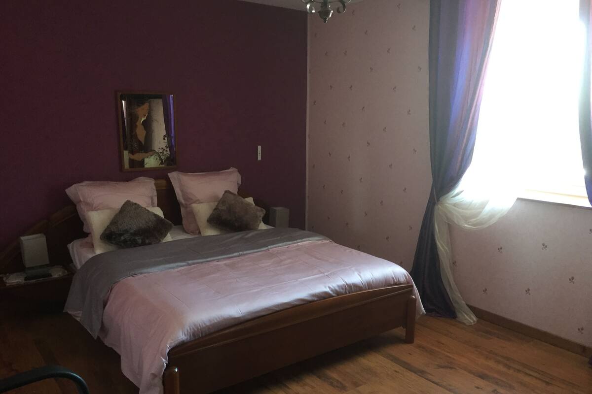 A bedroom with a wooden bed, purple walls, a nightstand, and a window with curtains.
