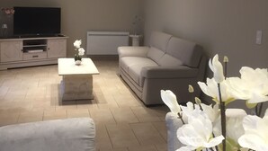 Un salón moderno con televisor de pantalla plana, un sofá gris, una mesa de centro y un florero con flores blancas.