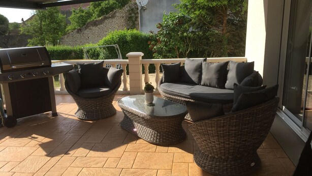 Un patio techado con mobiliario de mimbre, una parrilla y vistas a un muro de piedra y follaje.
