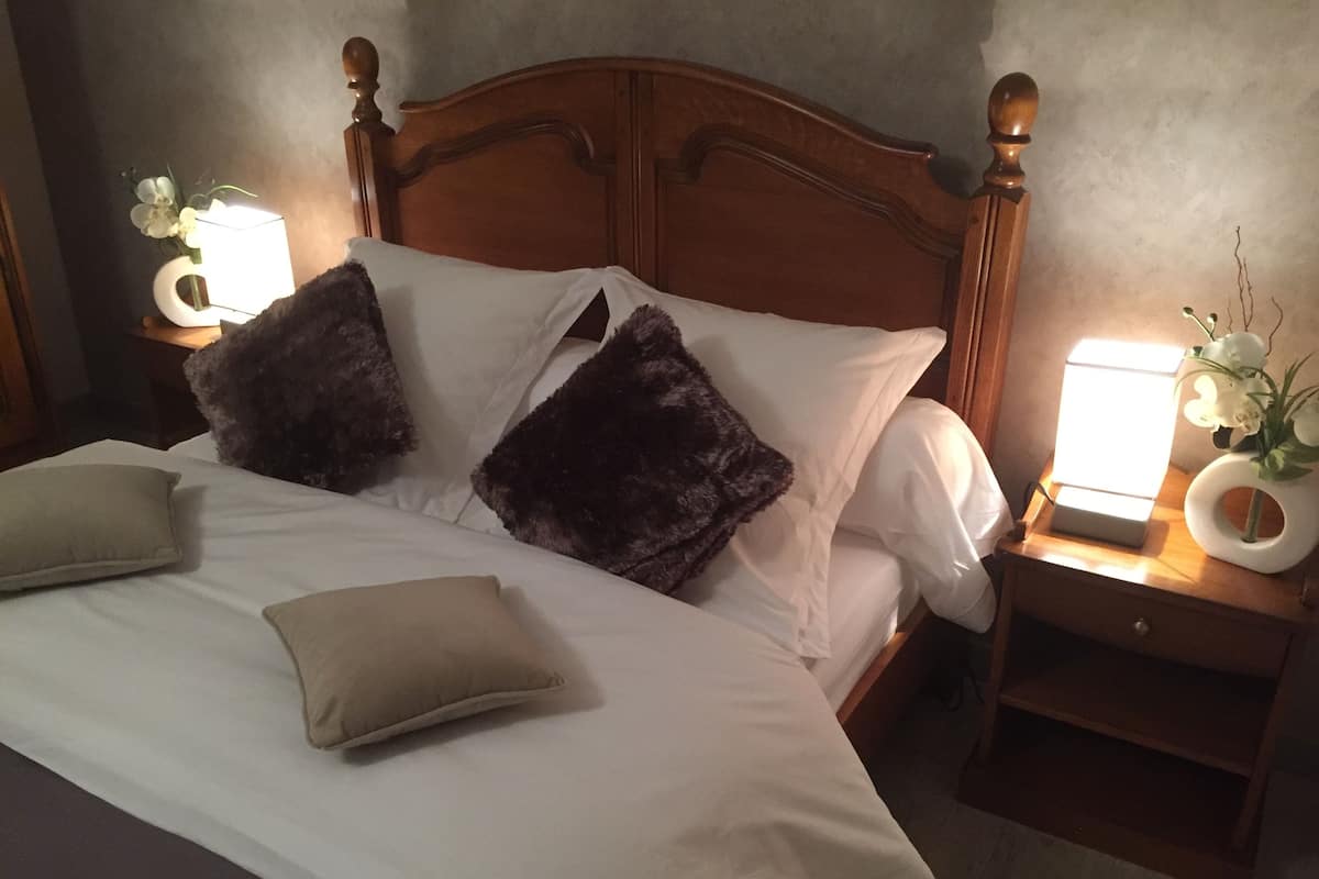 A neatly made bed with white linens and two pillows, a wooden headboard, and a bedside table with a lamp and a vase of flowers.