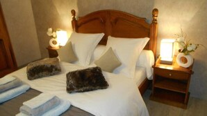 Una cama bien hecha con sábanas blancas, dos almohadas y dos mantas mullidas. Hay dos mesitas de noche con lámparas y un jarrón de flores.