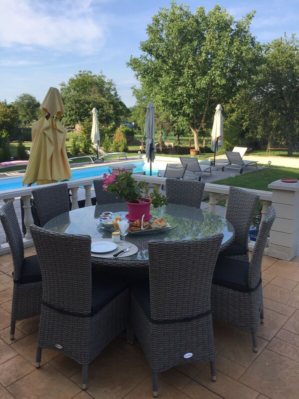 Uma área de refeições ao ar livre com cadeiras de vime e uma mesa de vidro, cercada por uma piscina e vegetação.