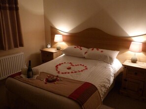 A hotel room with a bed, two bedside tables, a nightstand, a lamp, a bottle, and a heart-shaped decoration on the bed. - Dunamoy Cottages & Spa (Ballyclare)
