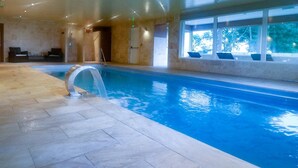 En indendørs swimmingpool med klart, blåt vand, flisegulv og et siddeområde med stole ved vinduet.