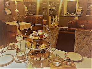 A three-tiered dessert stand with various pastries and a teapot on a table, surrounded by a cozy seating area with a sofa and a chair.