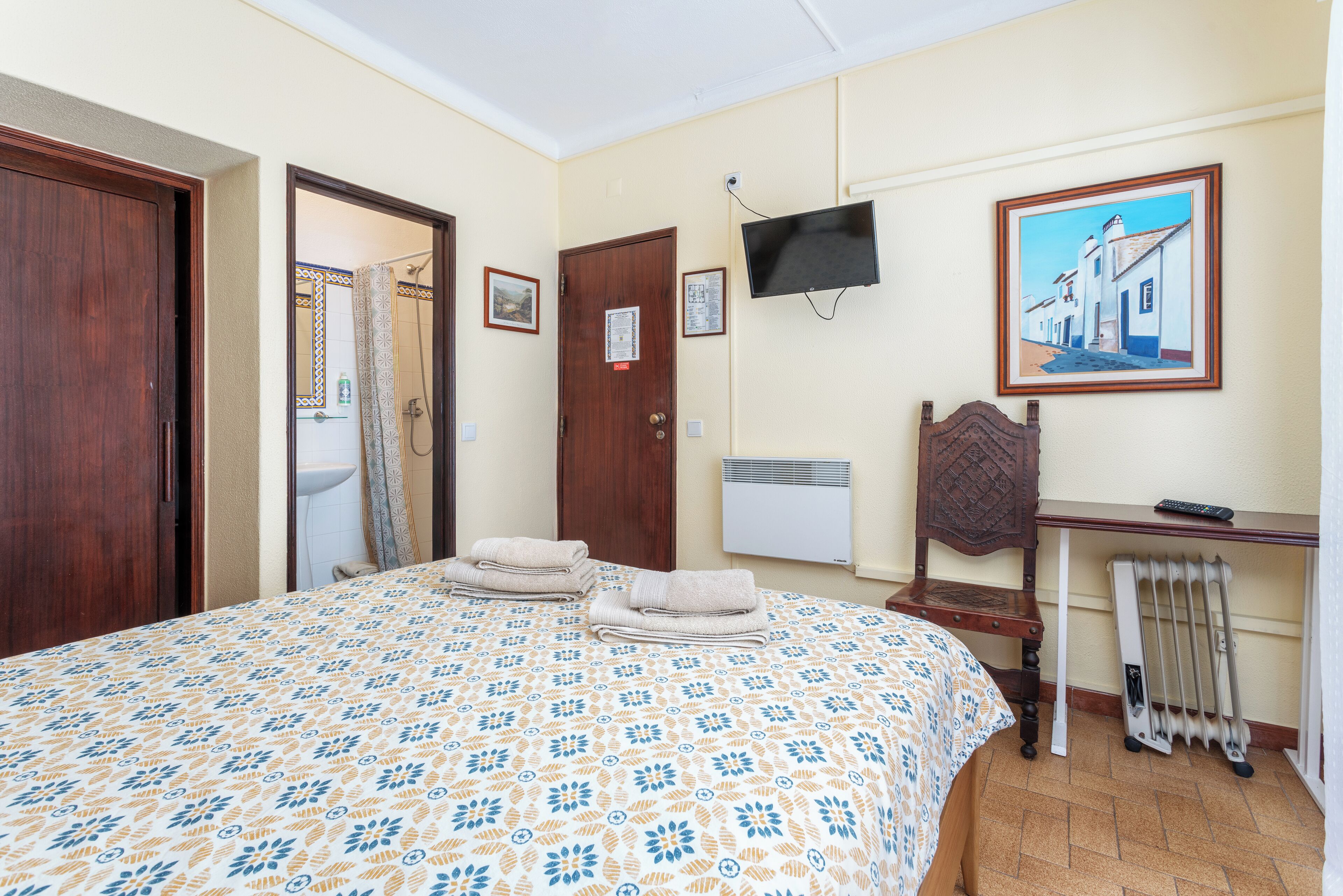 A hotel room with a bed, a desk, a chair, a radiator, a television, and a painting on the wall.