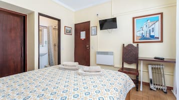 A hotel room with a bed, a desk, a chair, a radiator, a television, and a painting on the wall.